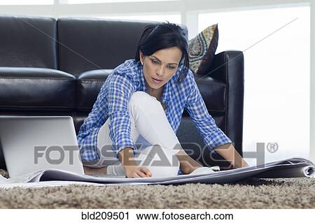 Stock Image - Hispanic woman examining blueprints in living room. Fotosearch