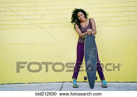 Stock Image - Hispanic woman holding skateboard on city street. Fotosearch