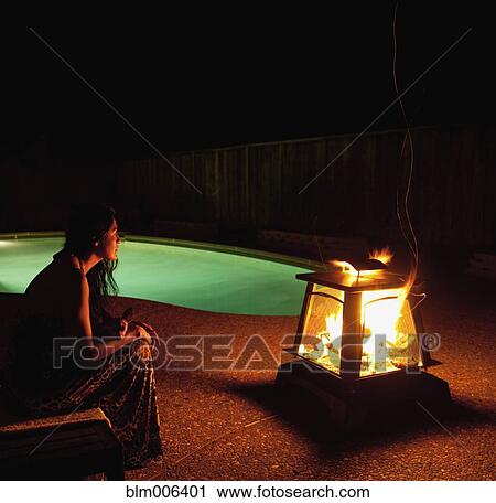 Stock Image - Hispanic woman sitting near fire pit near swimming pool. Fotosearch