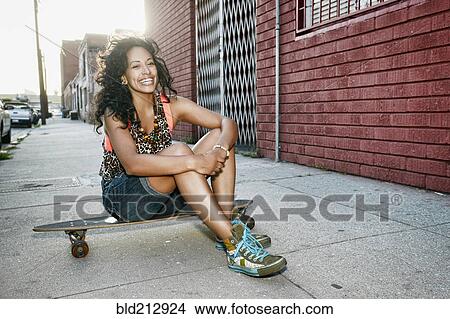 Picture - Hispanic woman sitting on skateboard on city street. Fotosearch