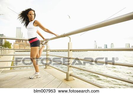 Stock Image - Hispanic woman stretching against railing at waterfront. Fotosearch