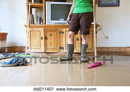 Stock Photo - Hispanic woman sweeping water out of flooded house. Fotosearch