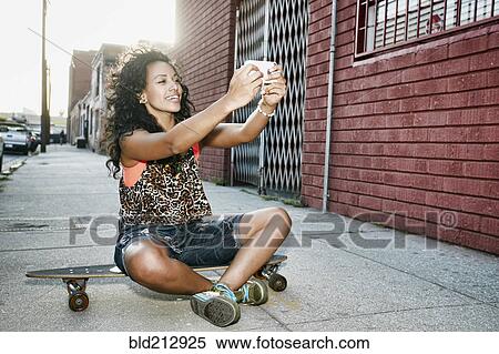 Stock Photography - Hispanic woman taking cell phone picture on skateboard on city street. Fotosearch