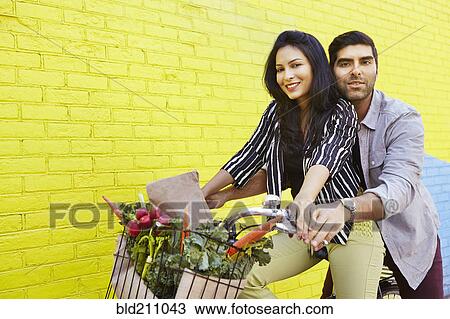 Stock Image - Indian couple riding bicycle along yellow wall. Fotosearch