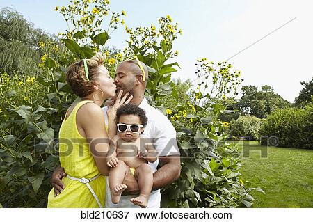 Stock Photo - Kissing parents holding baby girl in park. Fotosearch
