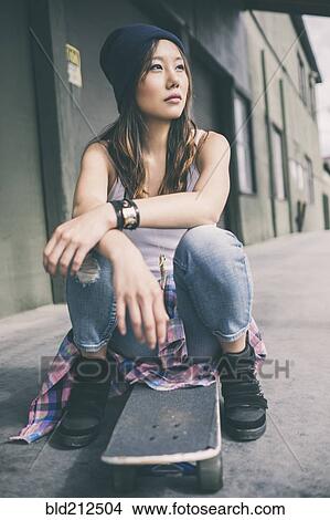 Picture - Korean woman sitting on skateboard on city street. Fotosearch