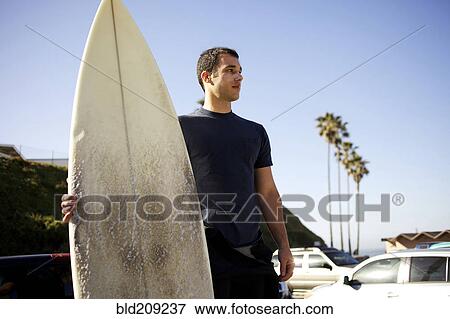 Stock Photo - Man holding surfboard in parking lot. Fotosearch