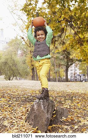 Stock Image - Mixed race boy holding pumpkin overhead in park. Fotosearch