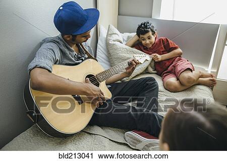 Stock Image - Mixed race boy watching brother playing guitar in bedroom. Fotosearch