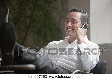 Stock Image - Mixed race businessman with feet on desk using cell phone. Fotosearch