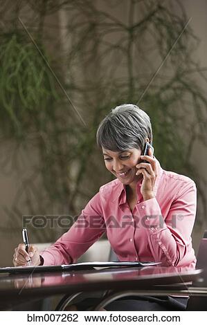 Stock Image - Mixed race businesswoman writing and using cell phone. Fotosearch