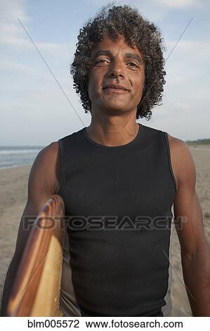 Stock Image - Mixed race man holding surfboard on beach. Fotosearch