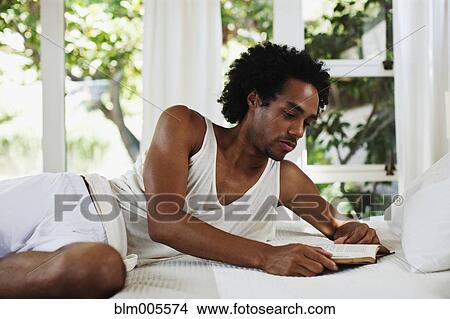 Picture - Mixed race man laying on bed reading. Fotosearch