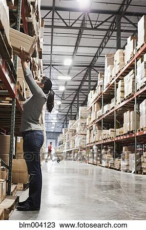 Stock Image - Mixed race woman reaching for box in warehouse. Fotosearch