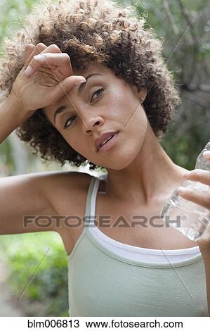 Stock Image - Mixed race woman wiping forehead and drinking water. Fotosearch
