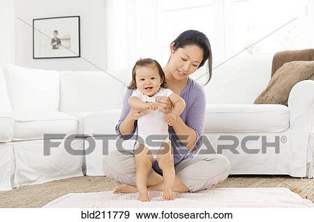 Stock Photo - Mother helping baby girl stand in living room. Fotosearch