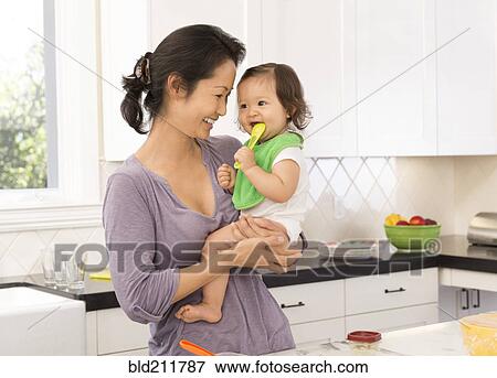 Stock Photo - Mother holding baby girl in kitchen. Fotosearch