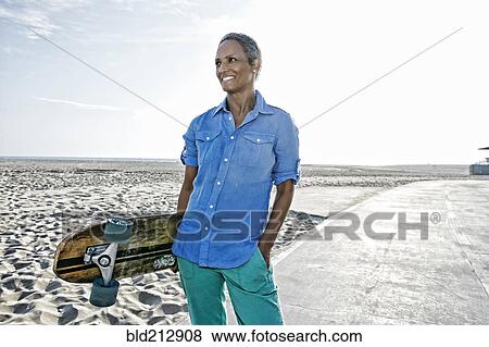 Stock Photo - Older Black woman holding skateboard on beach. Fotosearch