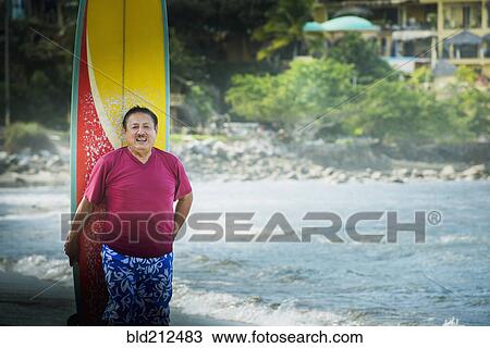 Stock Image - Older Hispanic man with surfboard on beach. Fotosearch
