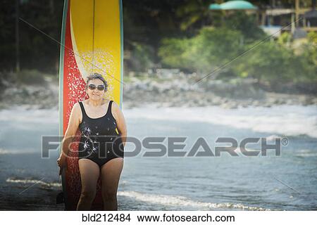 Picture - Older Hispanic woman with surfboard on beach. Fotosearch