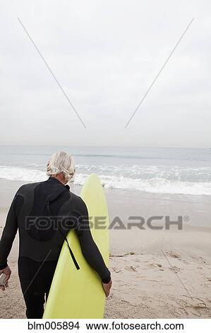 Picture - Senior Hispanic man carrying surfboard. Fotosearch