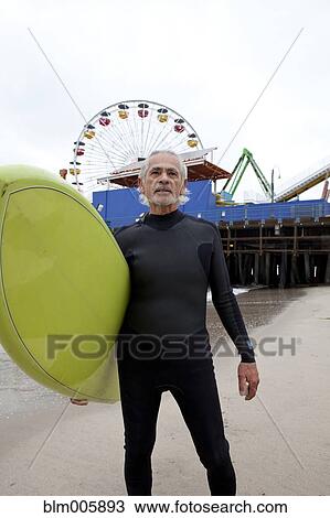 Stock Image - Senior Hispanic man carrying surfboard. Fotosearch