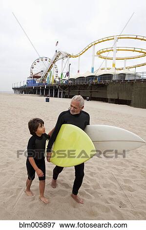 Stock Photo - Senior Hispanic man carrying surfboards with grandson. Fotosearch