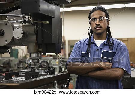 Stock Photography - Serious Hispanic man with arms crossed in machine shop. Fotosearch