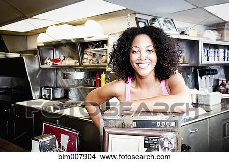Picture - Smiling African American woman leaning on cash register in diner. Fotosearch