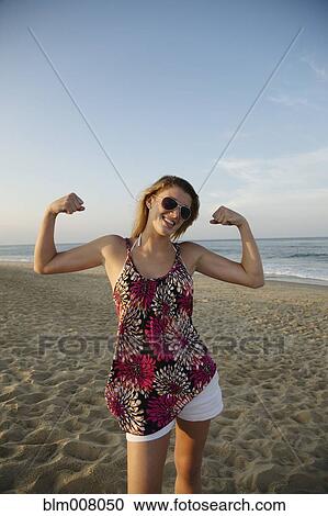 Stock Image - Smiling Caucasian teenage girl flexing biceps on beach. Fotosearch