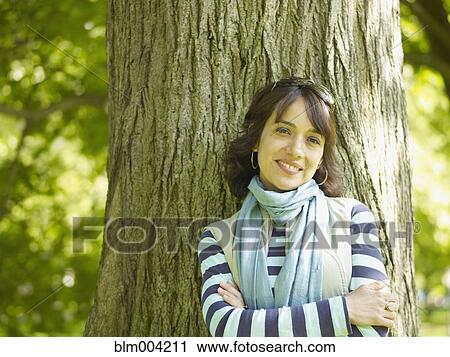 Stock Image - Smiling Hispanic woman leaning against tree with arms crossed. Fotosearch