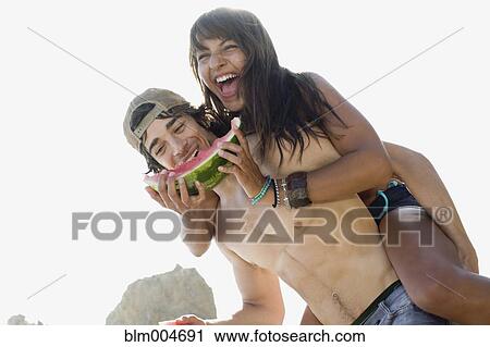 Stock Image - Woman eating watermelon getting piggy back ride from boyfriend. Fotosearch