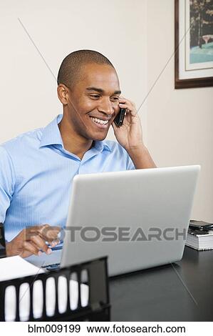 Stock Photo - African American talking on cell phone at desk. Fotosearch