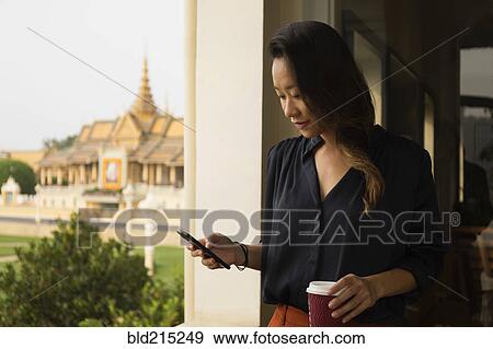 Stock Photo - Asian businesswoman using cell phone at window, Phnom Penh, Cambodia. Fotosearch