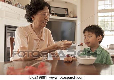Stock Image - Asian grandmother feeding grandson at table. Fotosearch