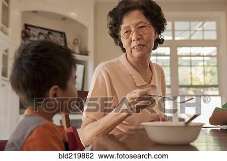 Stock Image - Asian grandmother feeding grandson at table. Fotosearch