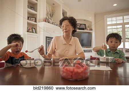 Stock Image - Asian grandmother feeding grandsons at table. Fotosearch