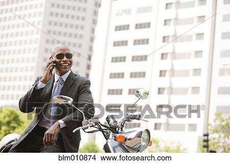 Picture - Black businessman sitting on scooter talking on cell phone. Fotosearch