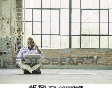 Stock Photo - Black businessman using laptop and cell phone on warehouse floor. Fotosearch