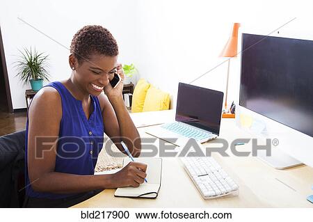 Stock Image - Black businesswoman talking on cell phone at office desk. Fotosearch