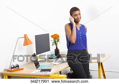 Stock Image - Black businesswoman talking on cell phone at office desk. Fotosearch