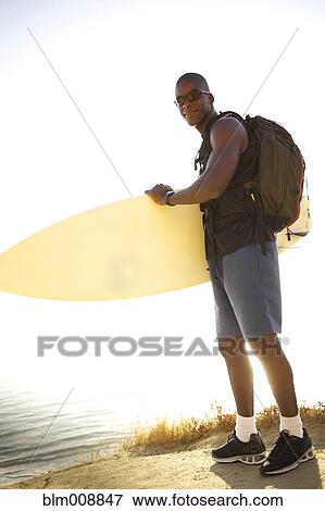Stock Photo - Black man carrying surfboard near ocean. Fotosearch