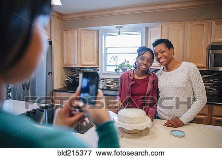 Stock Photo - Black woman photographing sister and mother in kitchen. Fotosearch