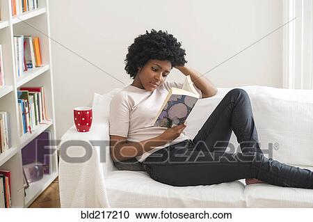 Stock Image - Black woman reading book on sofa. Fotosearch