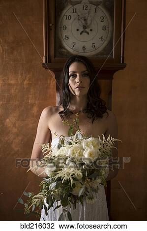 Stock Image - Bride holding bouquet of flowers under grandfather clock. Fotosearch
