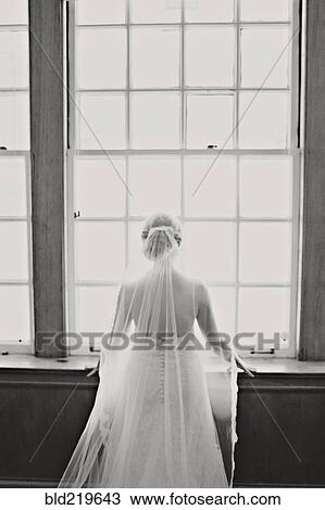 Stock Image - Bride in veil and wedding gown standing at window. Fotosearch