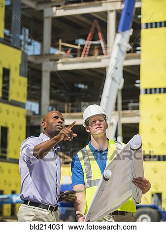 Stock Image - Businessman and construction worker reading blueprints at construction site. Fotosearch