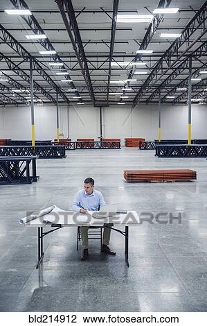 Stock Image - Caucasian architect reading blueprints in warehouse. Fotosearch