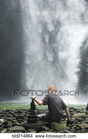 Picture - Caucasian boy stacking rocks near waterfall. Fotosearch