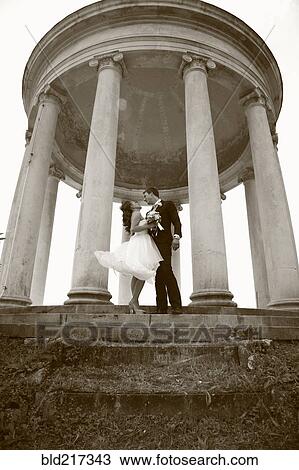 Stock Image - Caucasian bride and groom hugging under columns. Fotosearch